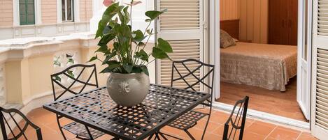 A balcony with a table and chairs, a potted plant, and a view of a bedroom with a bed and wardrobe.