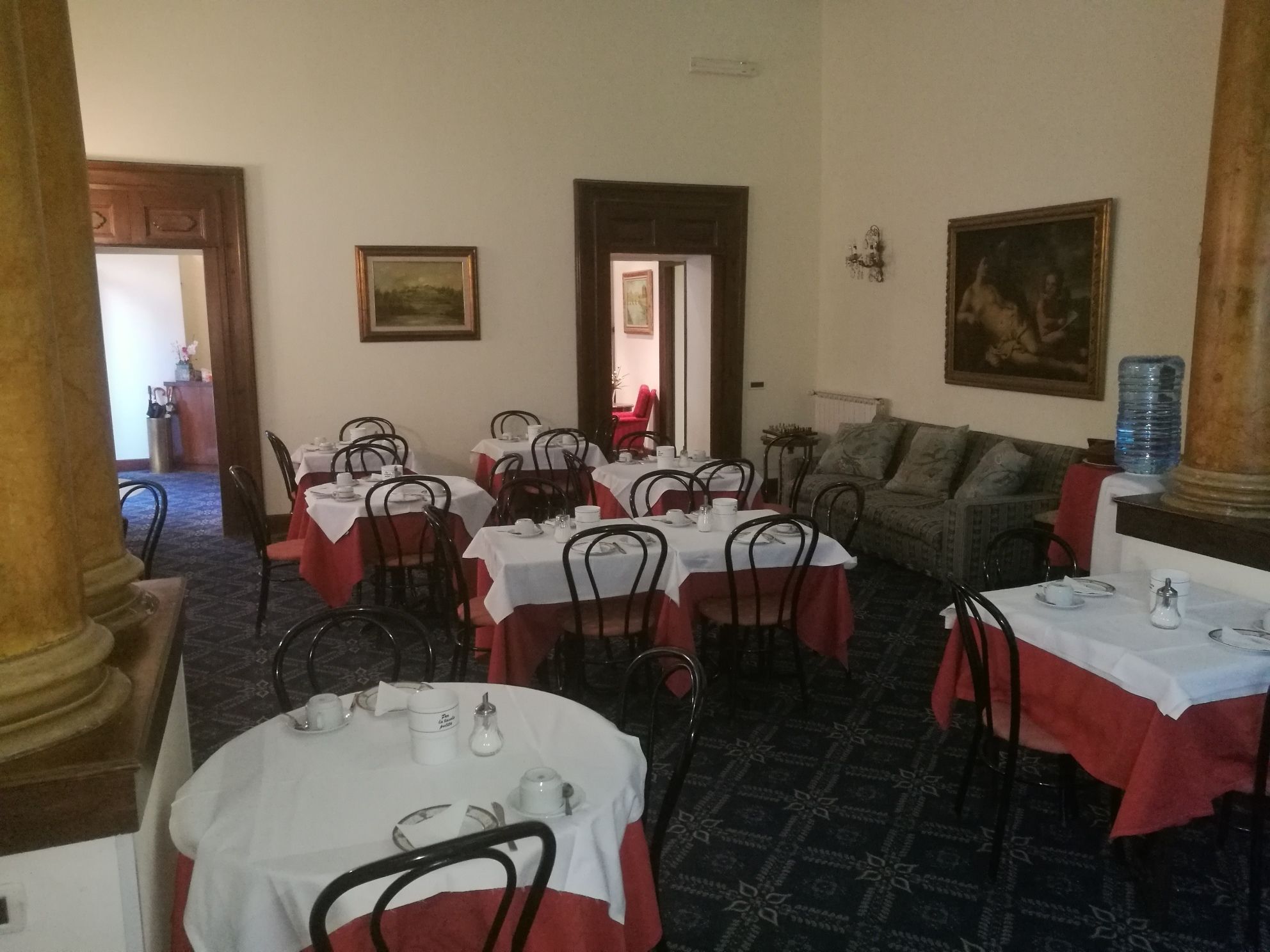 A dining area with round tables covered in white tablecloths and red table runners, surrounded by metal chairs. There is a sofa and a large water dispenser in the background.