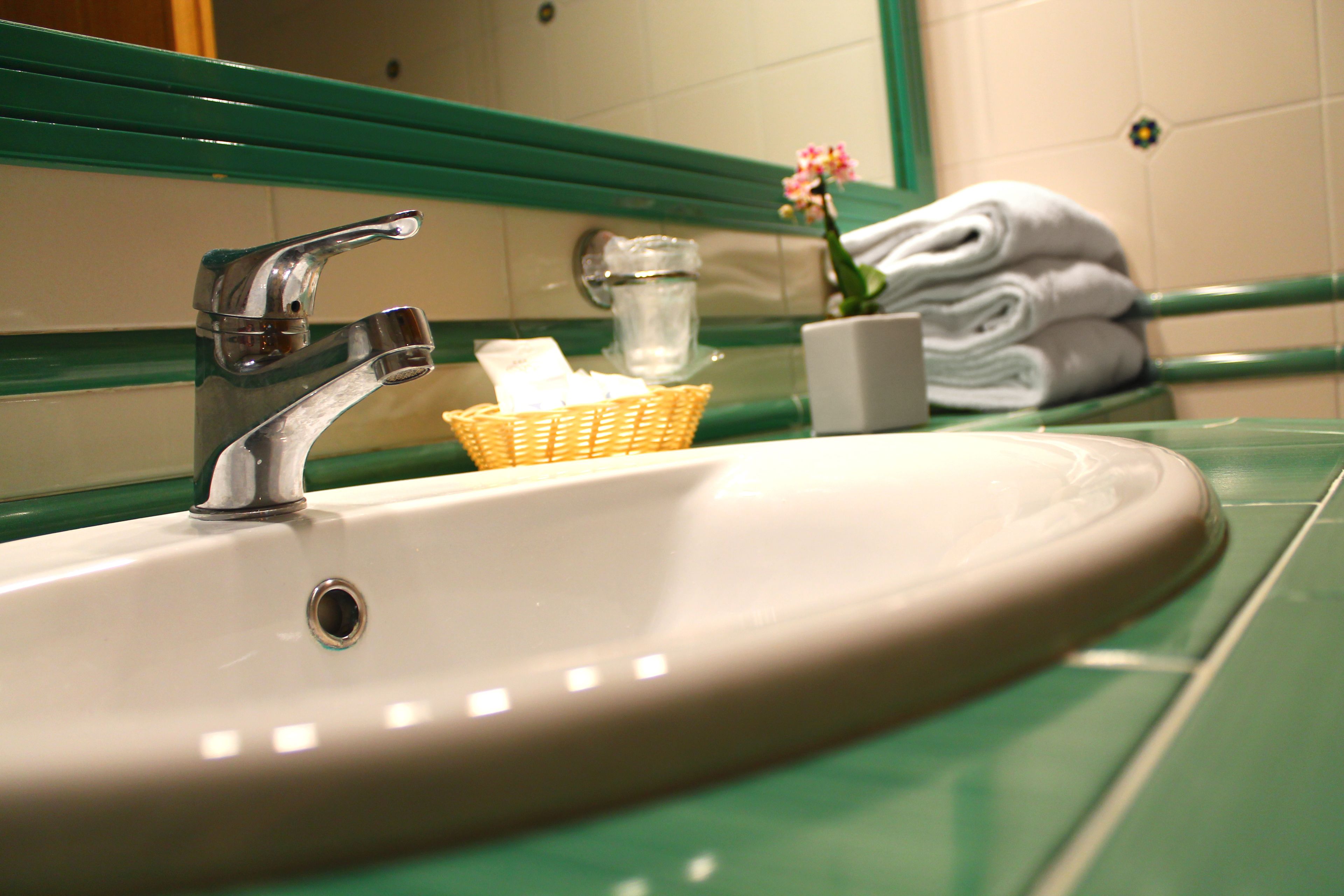 A bathroom sink with a chrome faucet, a glass pitcher, a wicker basket, a white cup, and a vase with pink flowers.