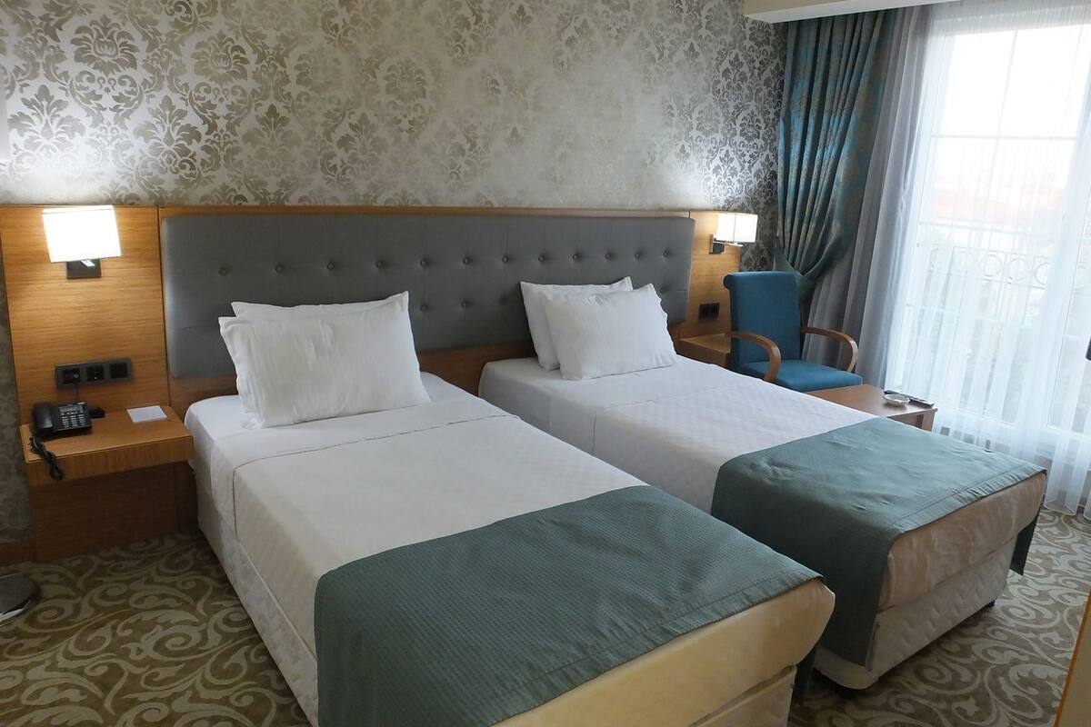 A hotel room with two beds, a headboard, bedside lamps, a chair, and a window with curtains.