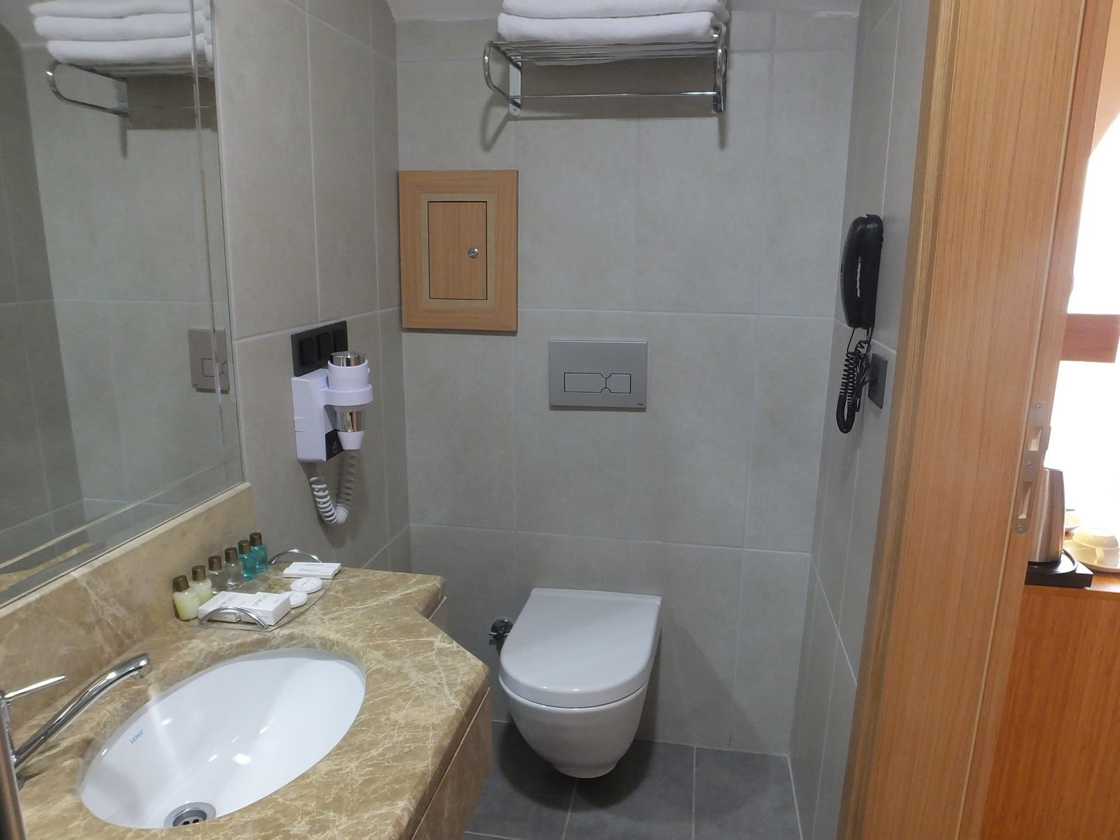 A bathroom with a sink, toilet, and a mirror. There are toiletries on the counter, a towel rack, and a phone on the wall.