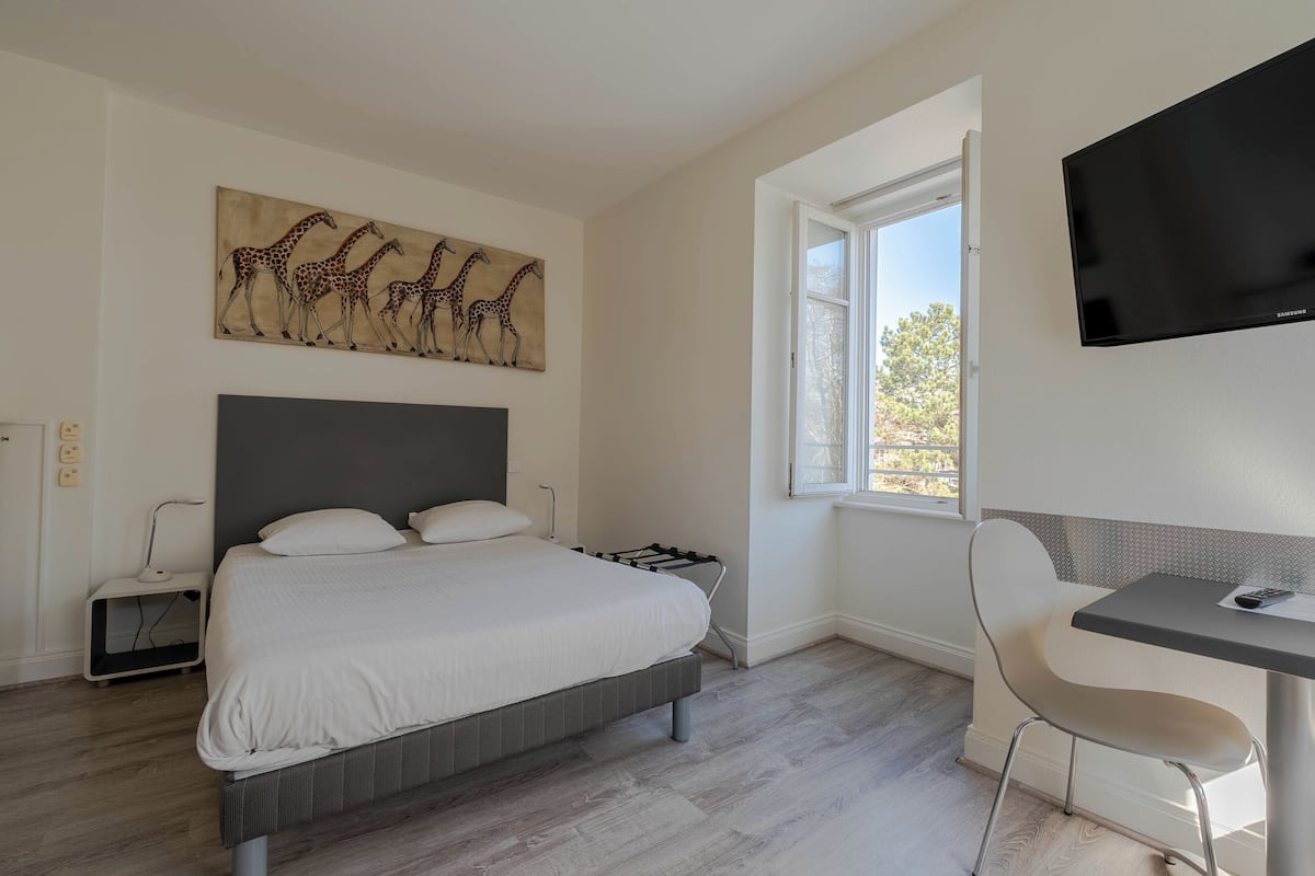 A bedroom with a bed, a desk, a chair, a TV, and a window with a view.