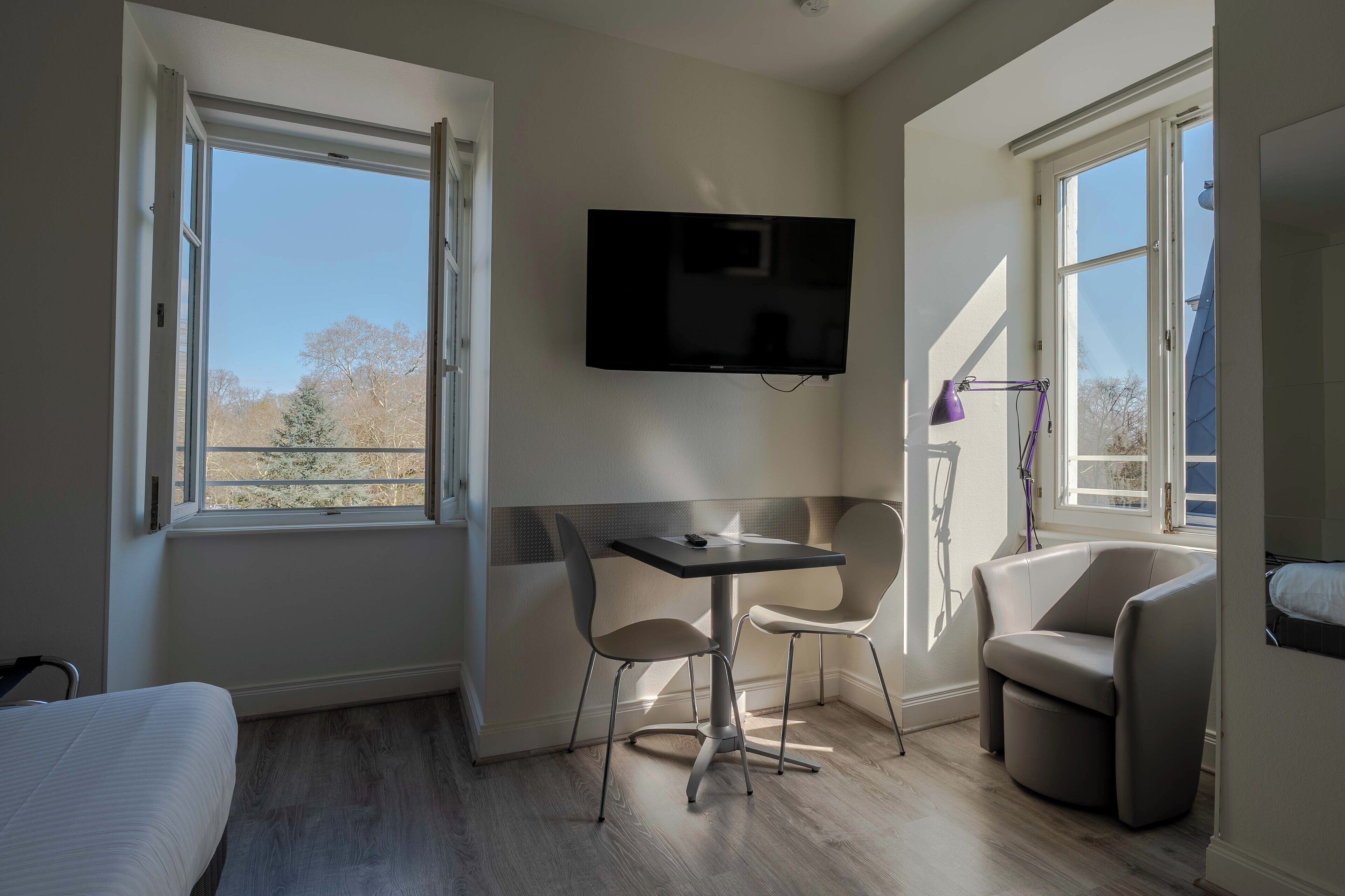 A small, modern room with a bed, a table, two chairs, a TV, and large windows.