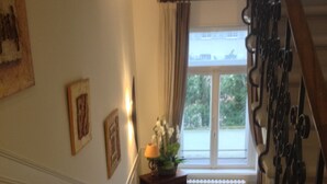 Una scala con ringhiera in legno, un tavolino con lampada e fiori, e una finestra con tende.
