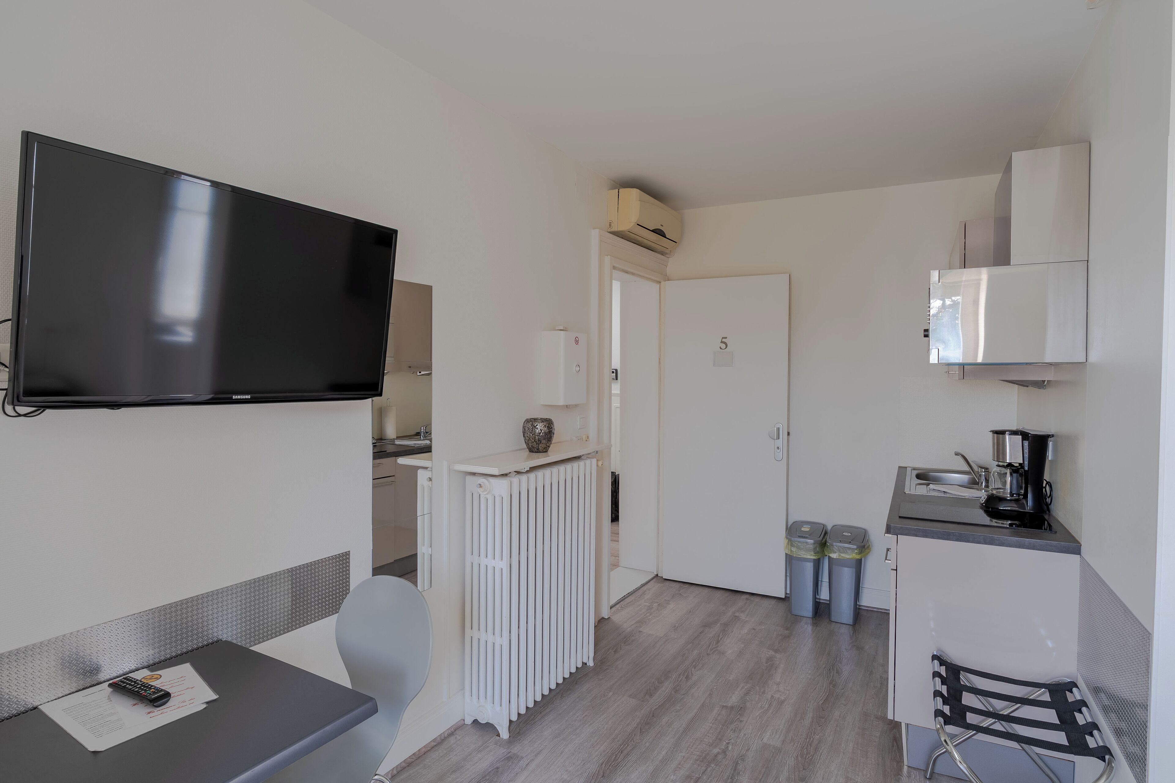 Une chambre d’hôtel compacte équipée d’une télévision, d’une petite table avec une télécommande, d’un radiateur, d’une kitchenette et d’une porte portant le numéro 5.