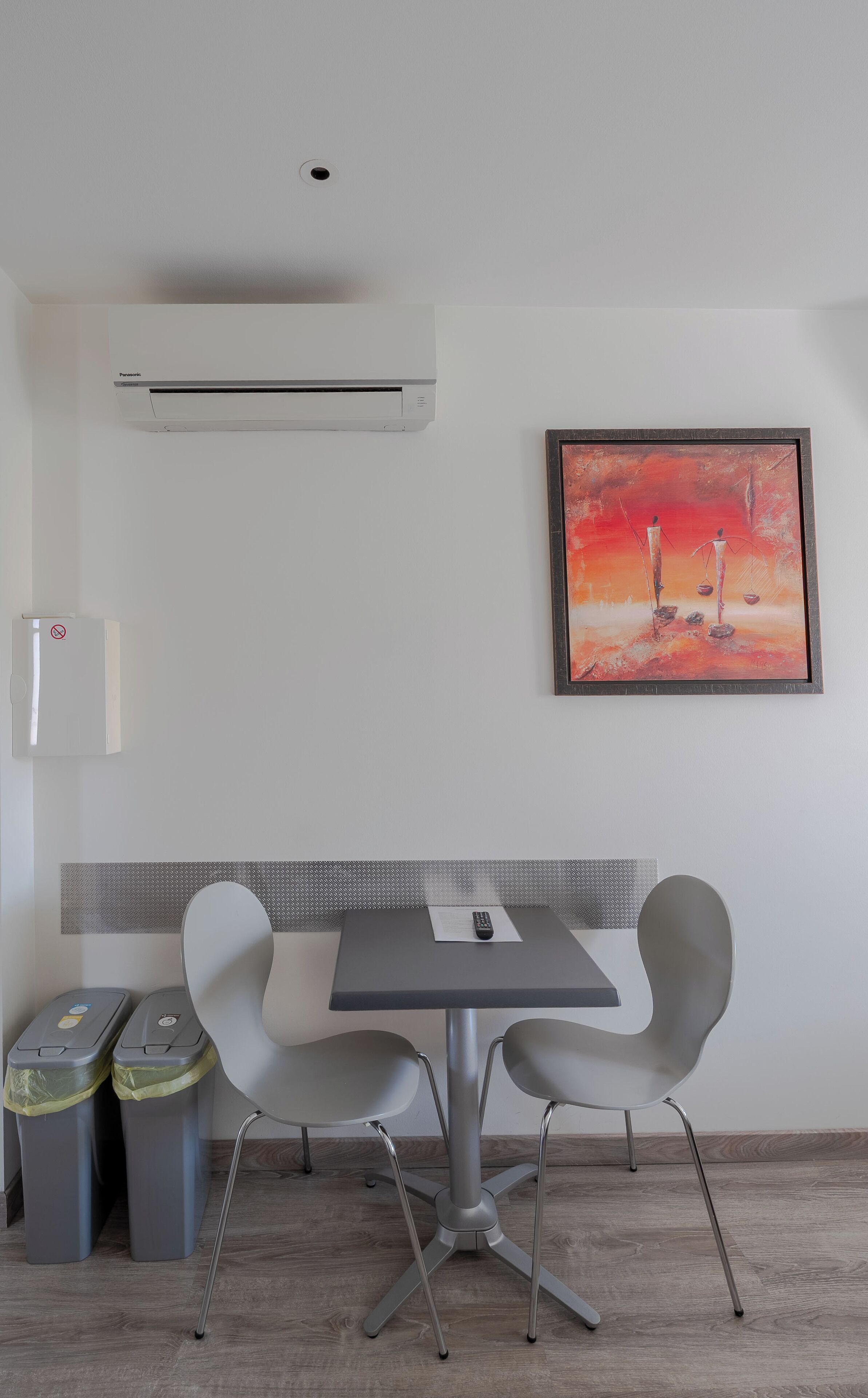 Una habitación moderna con una mesa gris y dos sillas, un cuadro enmarcado, un aire acondicionado blanco, un cubo de basura y un contenedor de reciclaje.