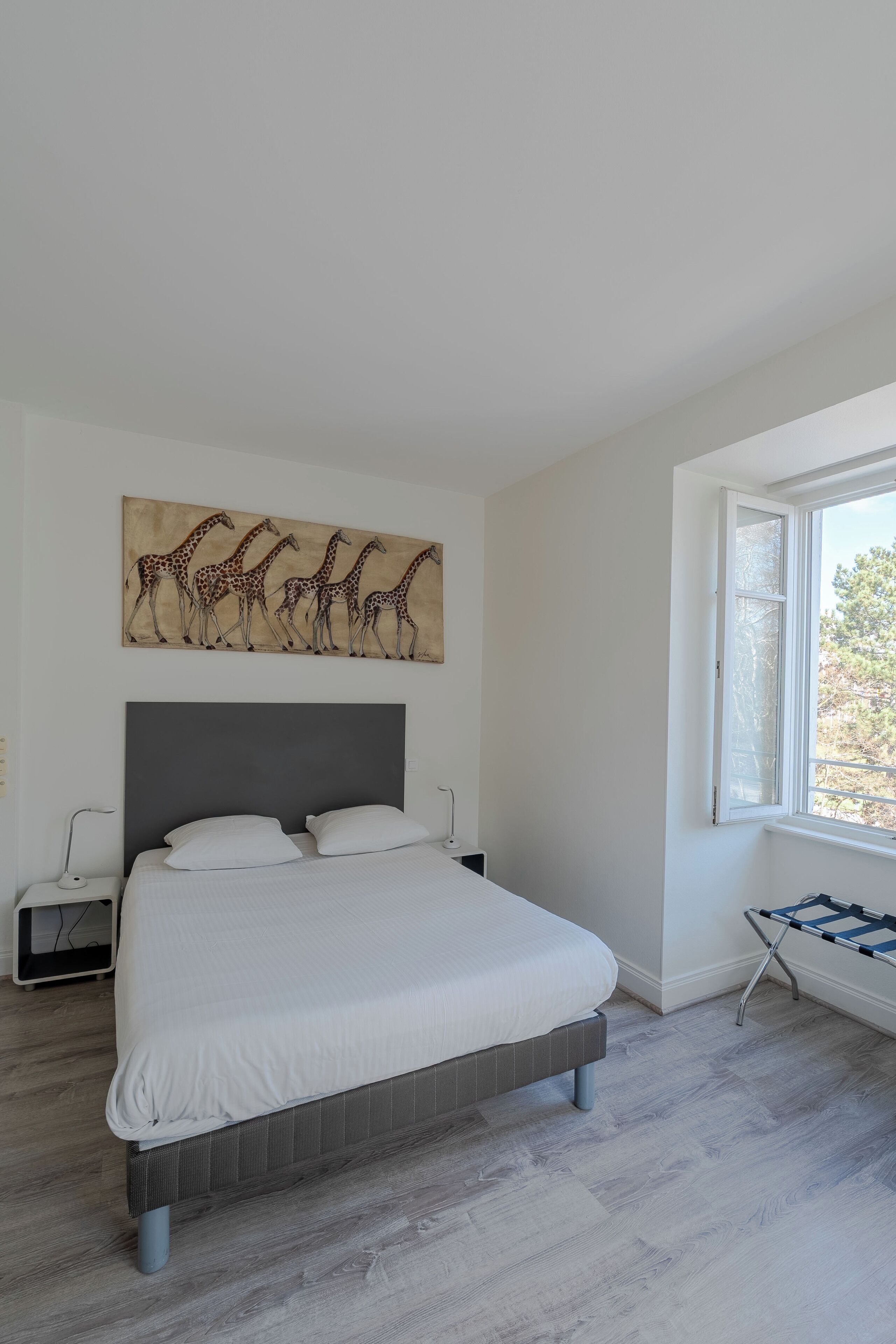 A bedroom with a large bed, a grey headboard, a giraffe painting on the wall, and a window with a view of trees.
