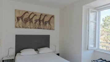 A bedroom with a large bed, a grey headboard, a giraffe painting on the wall, and a window with a view of trees.