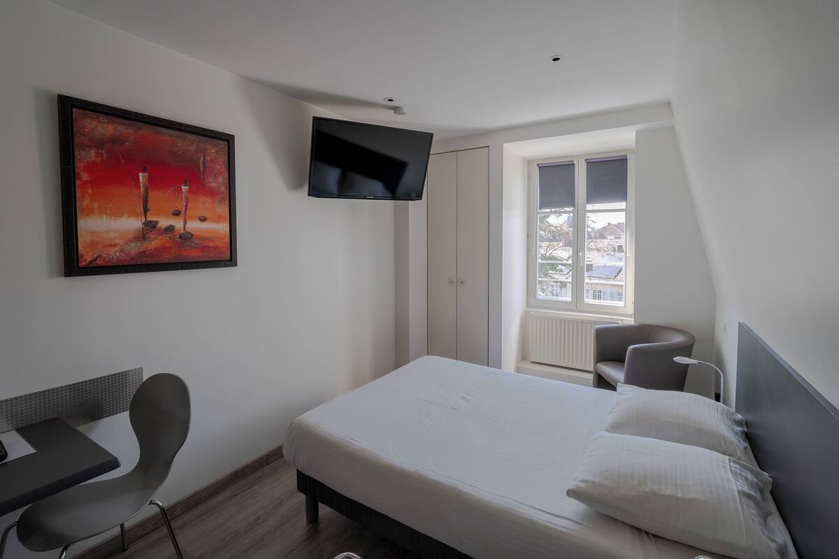 A bedroom with a bed, a desk, a chair, a TV, and a window with a view.