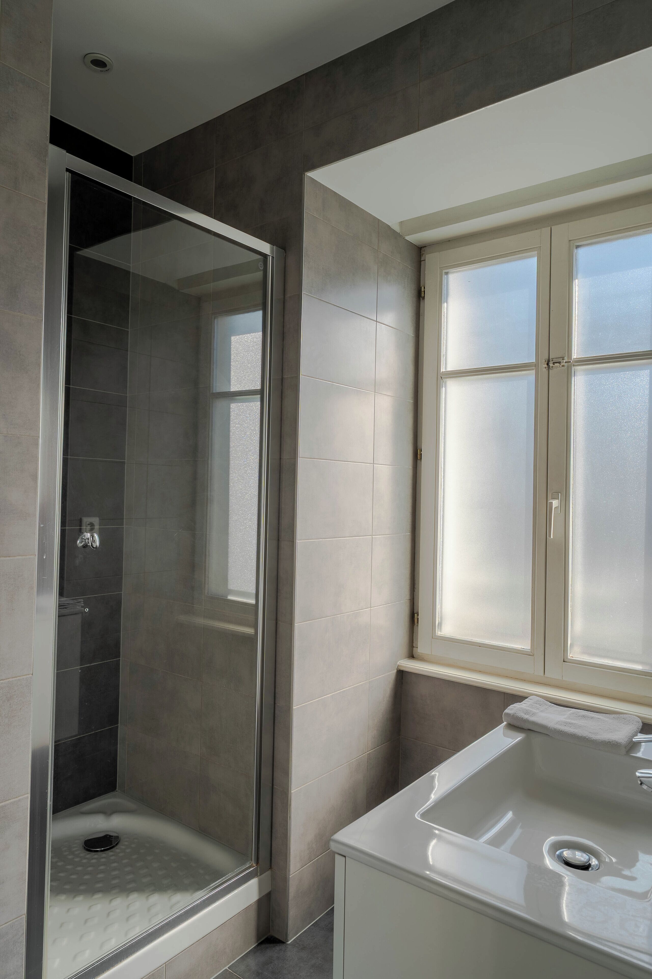 Ein modernes Badezimmer mit einer gläsernen Duschkabine, einem großen Fenster und einem weißen Waschbecken.