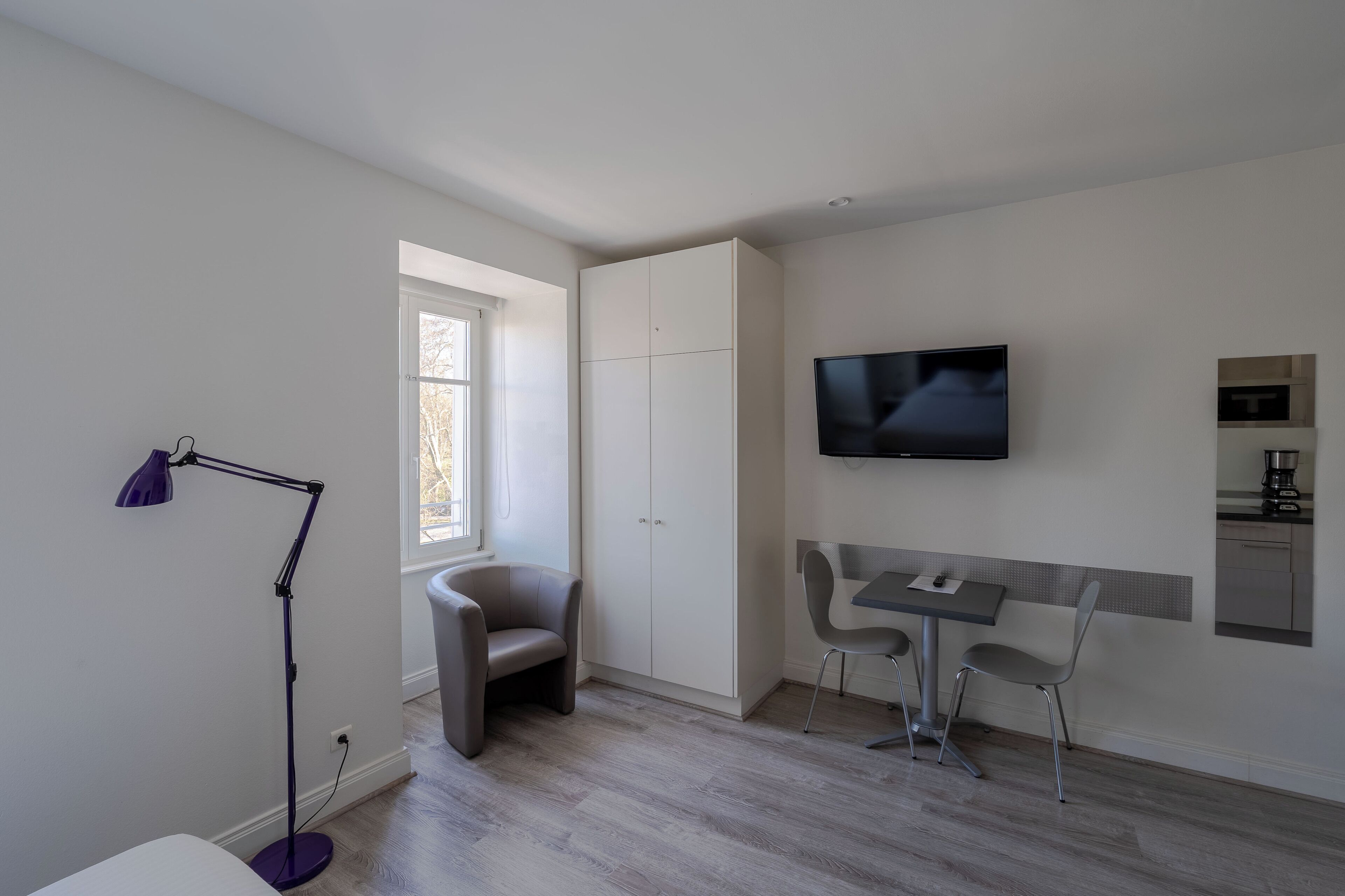 A modern, minimalist room with a dining area, a TV mounted on the wall, and a window providing natural light.