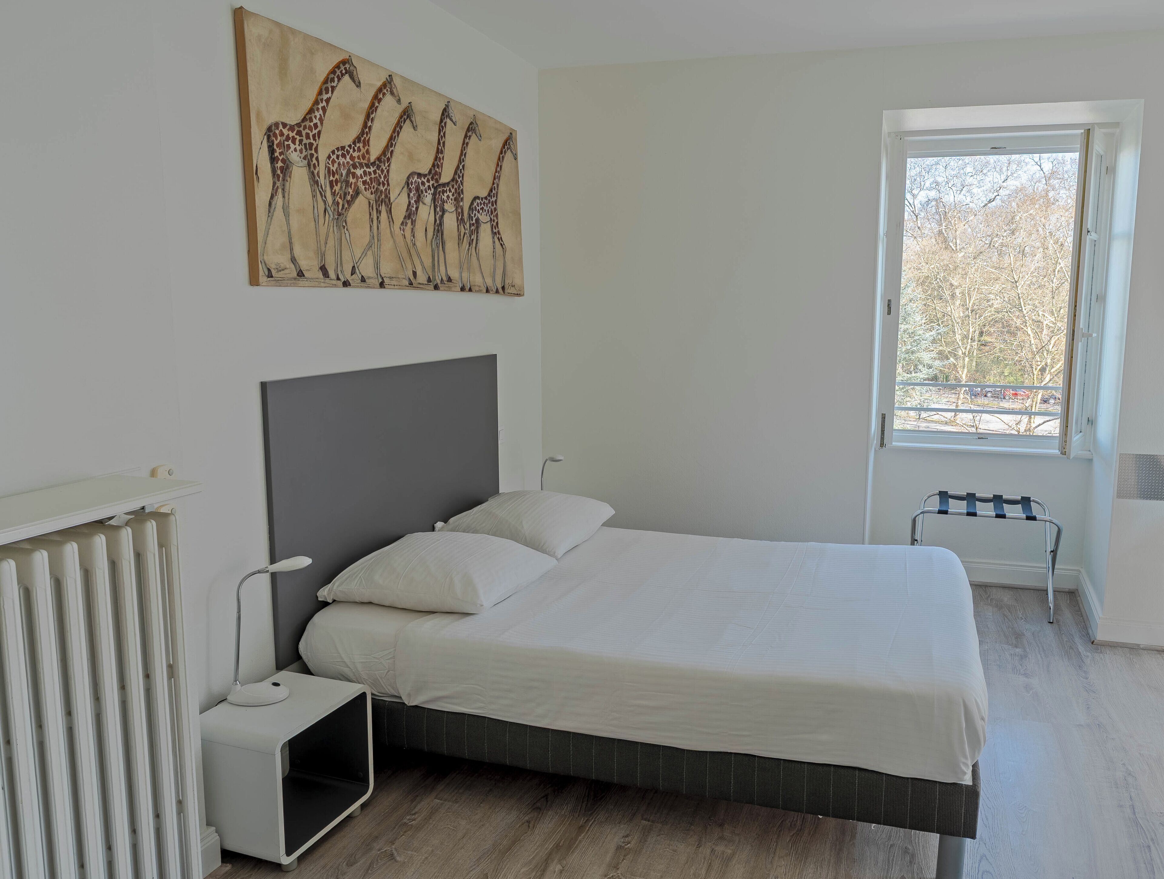 A bedroom with a bed, a radiator, a desk, a lamp, a window, and a giraffe painting on the wall.