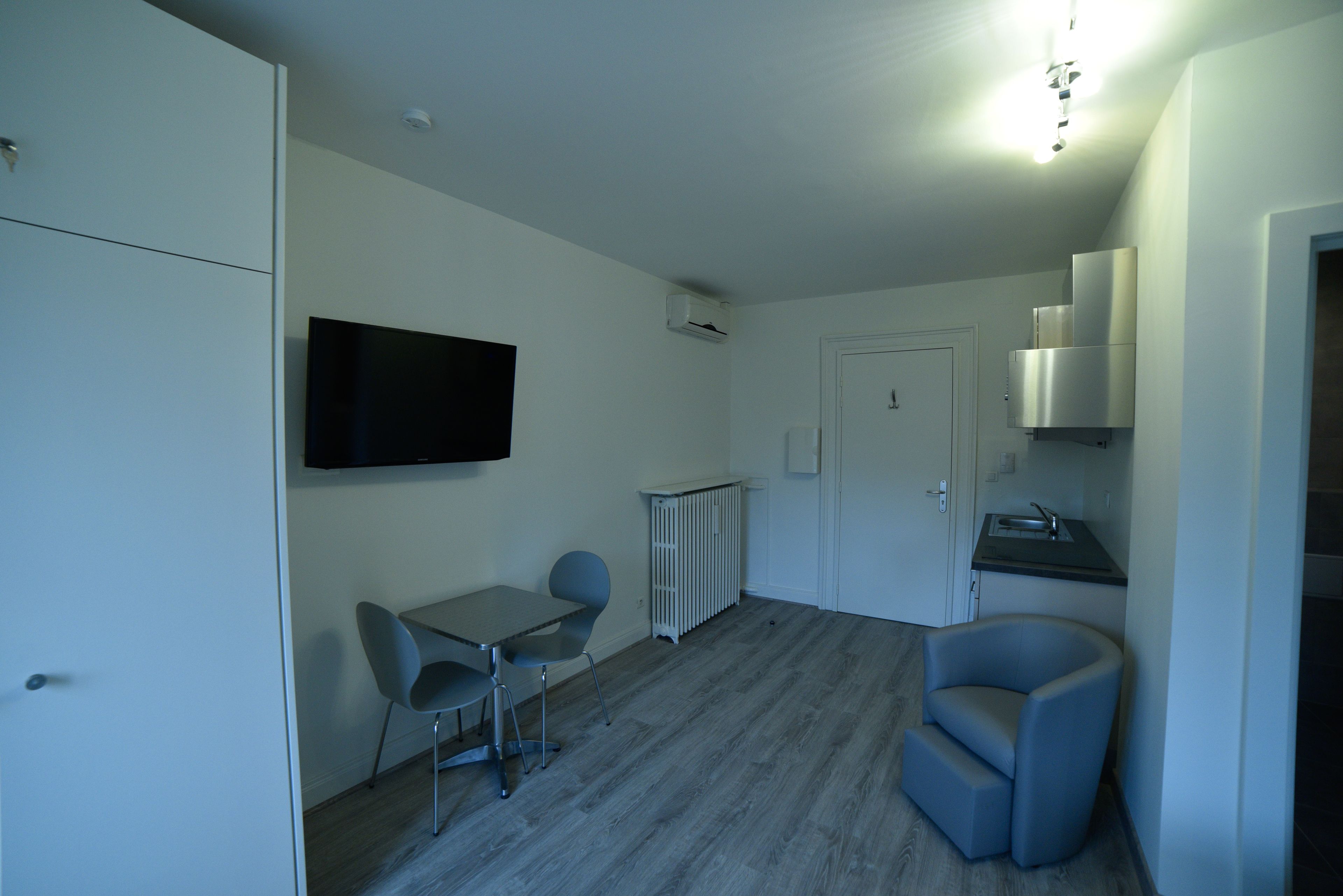 A compact room with a dining table, two chairs, a television, a kitchenette, and a door.