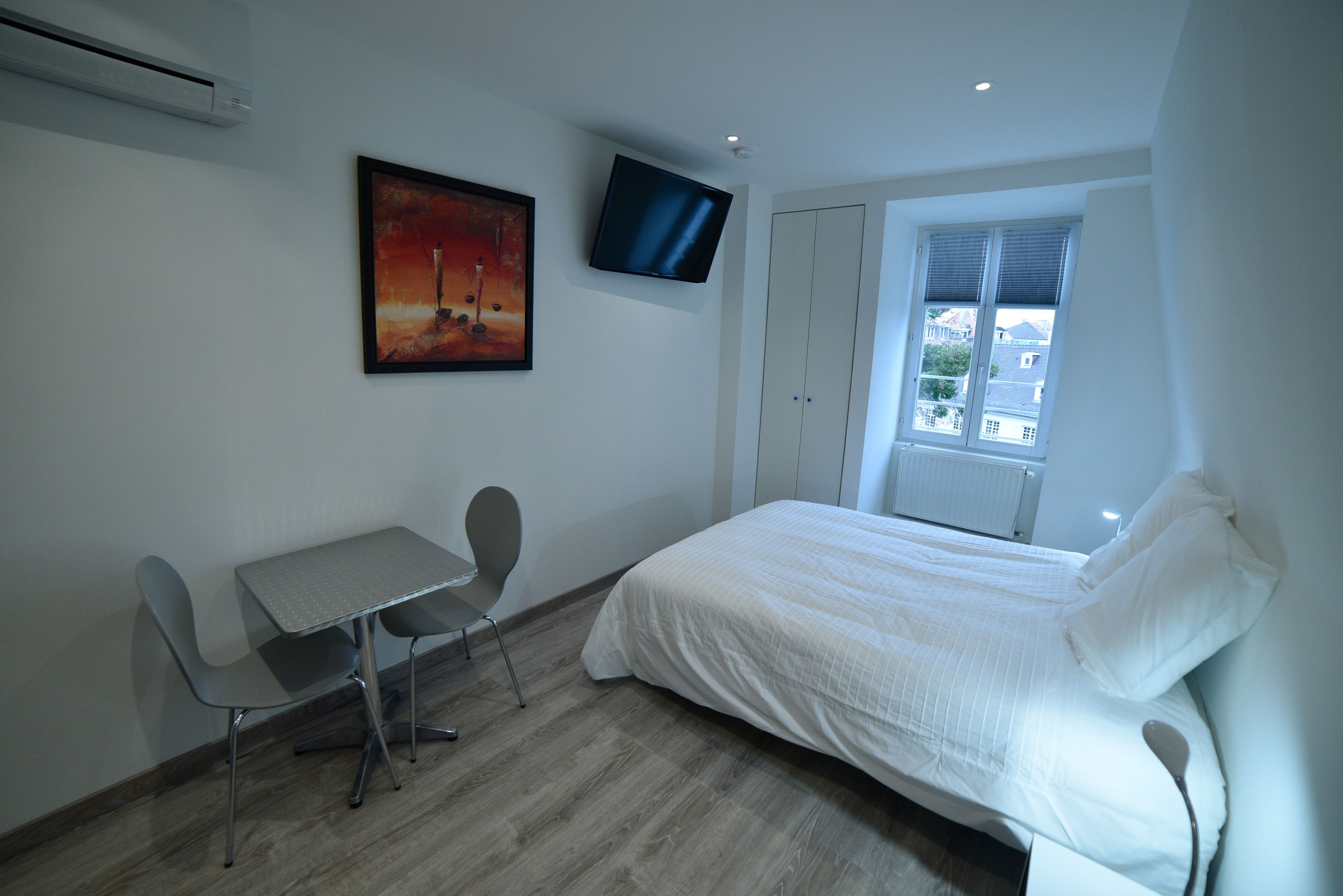 A bedroom with a bed, a desk, a chair, a TV, and a window.