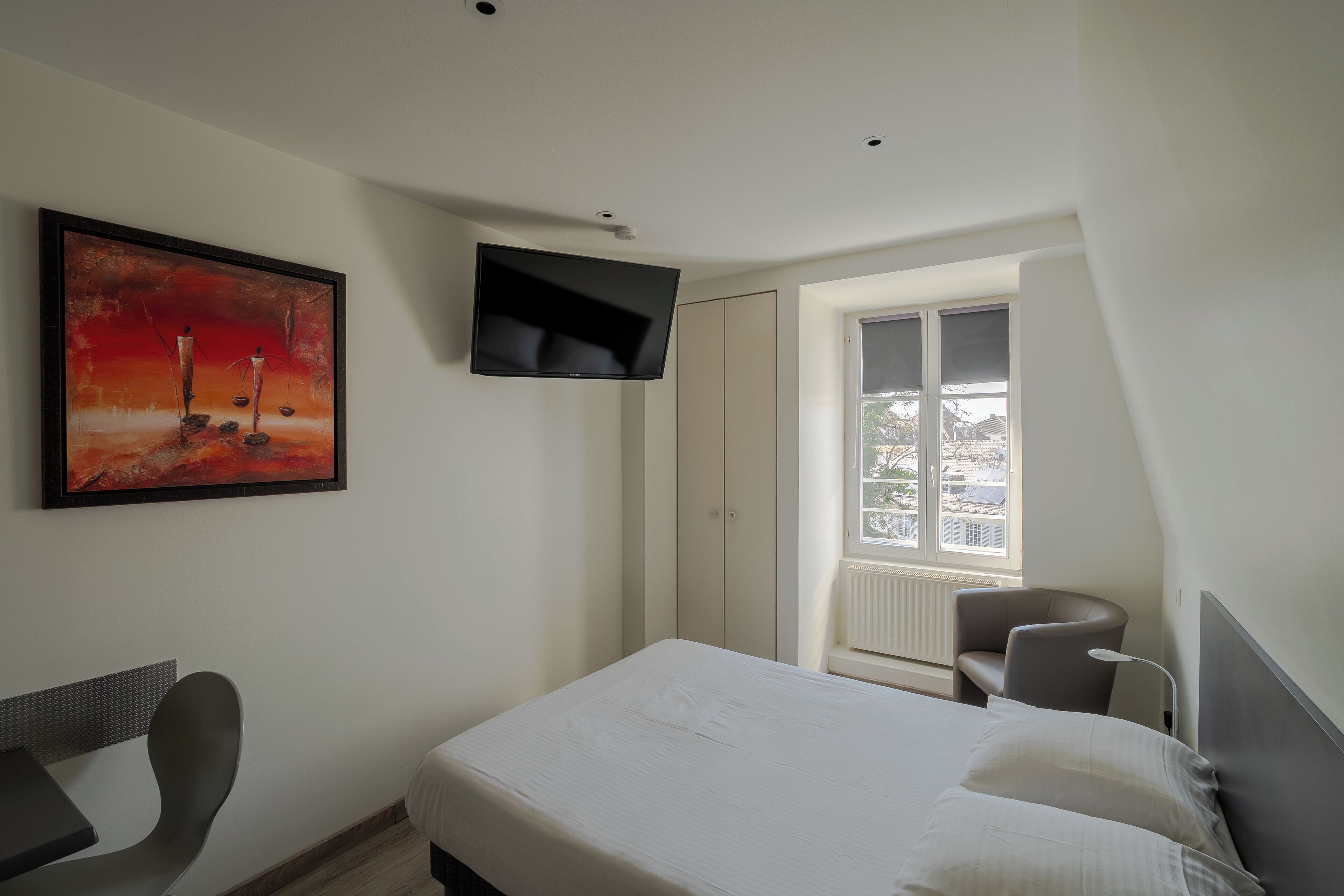 A hotel room with a bed, a TV mounted on the wall, a painting, a chair, and a window with a view.