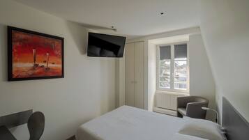 A hotel room with a bed, a TV mounted on the wall, a painting, a chair, and a window with a view.