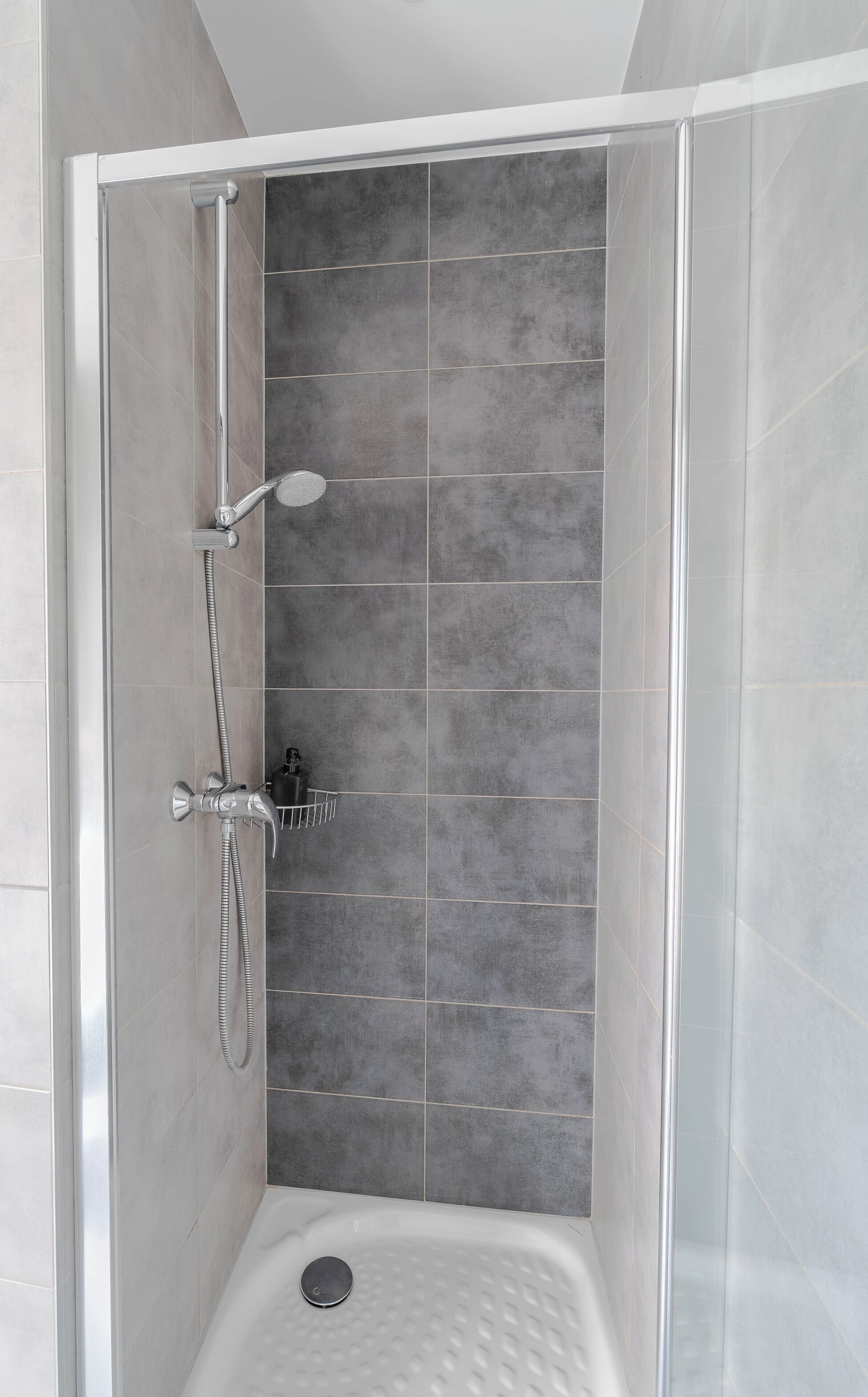 A walk-in shower with grey tiles, a handheld showerhead, and a wall-mounted faucet.