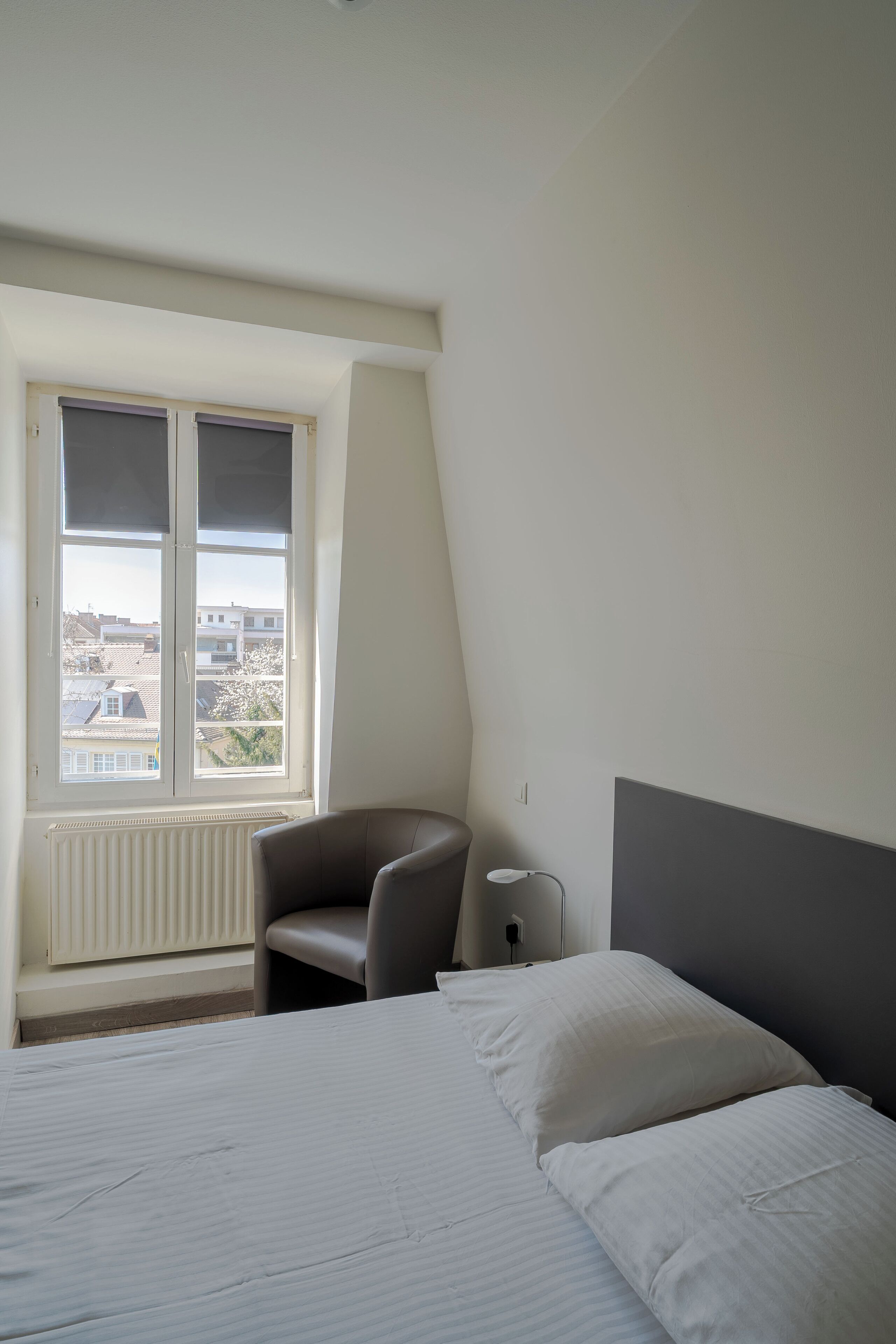 A bedroom with a bed, a chair, a desk, and a window with a view of buildings.