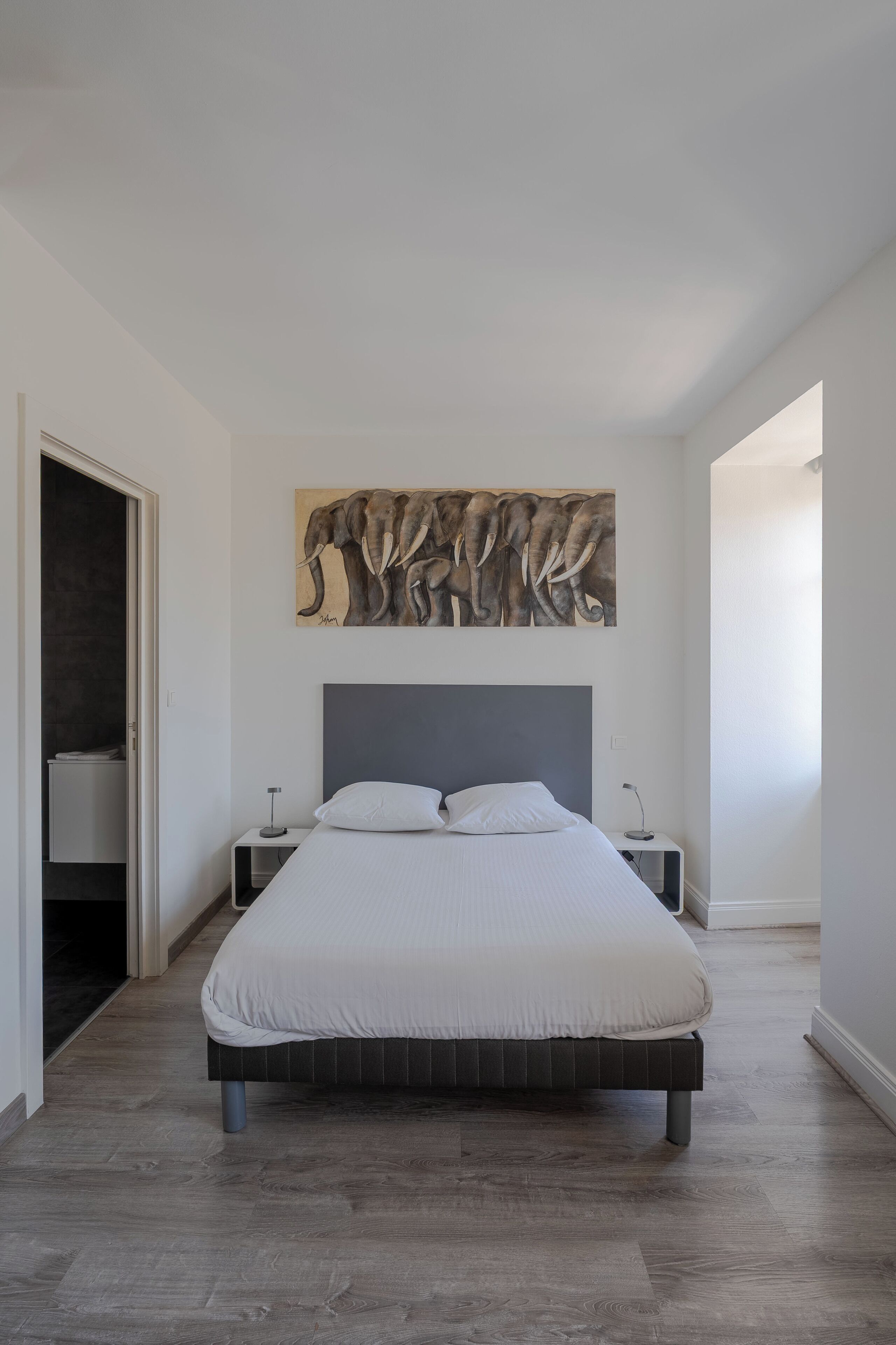 Une chambre moderne avec un grand lit, une tête de lit, une table de chevet, un tableau représentant un éléphant accroché au mur, et une porte donnant sur une autre pièce.