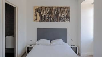 Une chambre moderne avec un grand lit, une tête de lit, une table de chevet, un tableau représentant un éléphant accroché au mur, et une porte donnant sur une autre pièce.