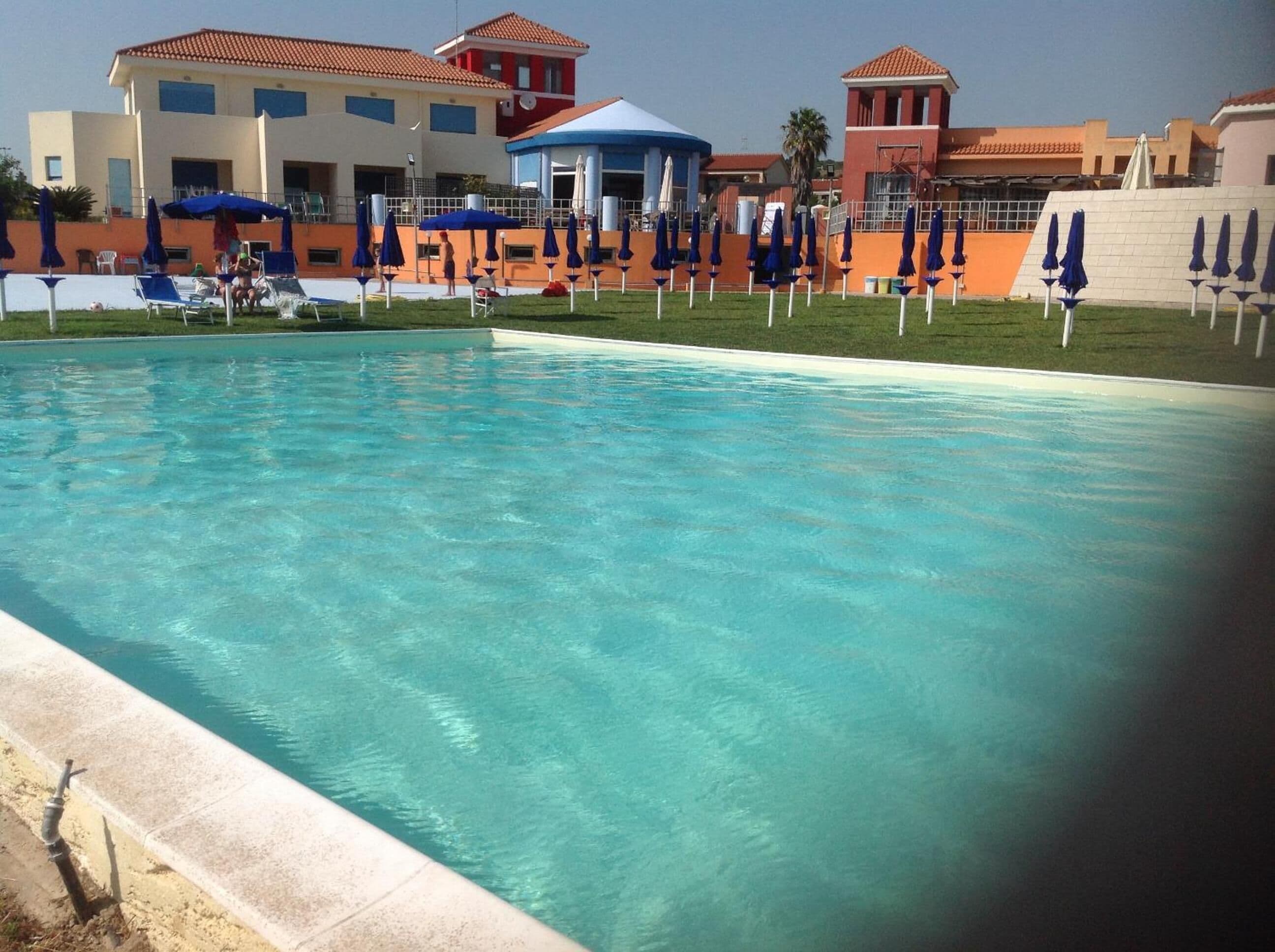 Une piscine aux eaux cristallines, bordée de chaises longues et d’ombrelles, et entourée d’immeubles aux toits recouverts de tuiles rouges.