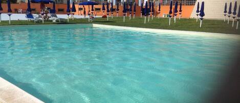 Une piscine aux eaux cristallines, bordée de chaises longues et d’ombrelles, et entourée d’immeubles aux toits recouverts de tuiles rouges.