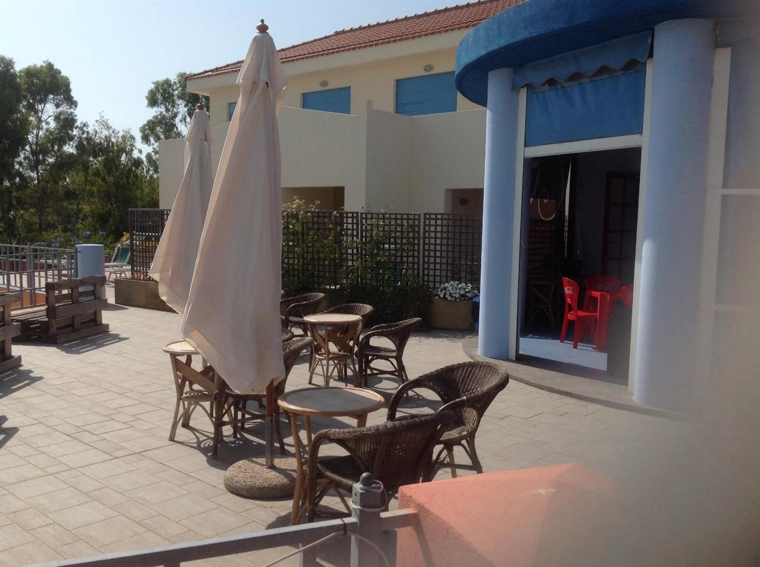 An outdoor patio area with wicker furniture, umbrellas, and a building entrance with red chairs inside.