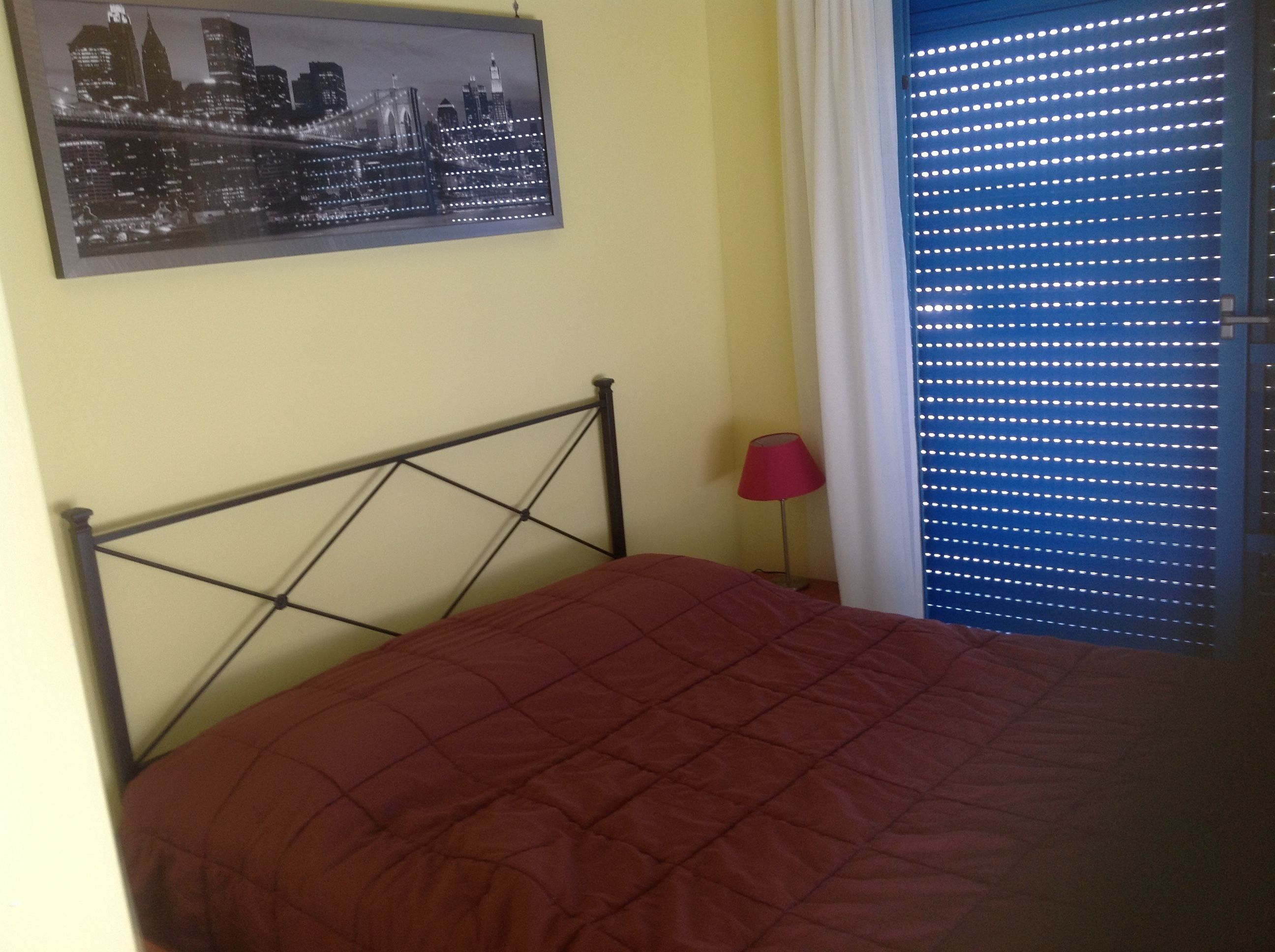 A bed with a metal frame and a red quilt, a red floor lamp, a cityscape painting on the wall, and a window with blue blinds.
