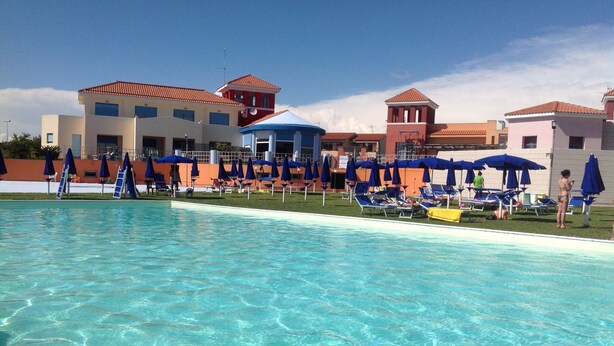 Una piscina con agua cristalina, rodeada de sillas de descanso y sombrillas, con edificios de techo rojo al fondo.