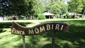 A wooden sign with the text "Finca Mombiri" in a rustic style, set against a backdrop of a well-maintained lawn and trees, with buildings in the distance. - Finca Mombiri (Esquina)