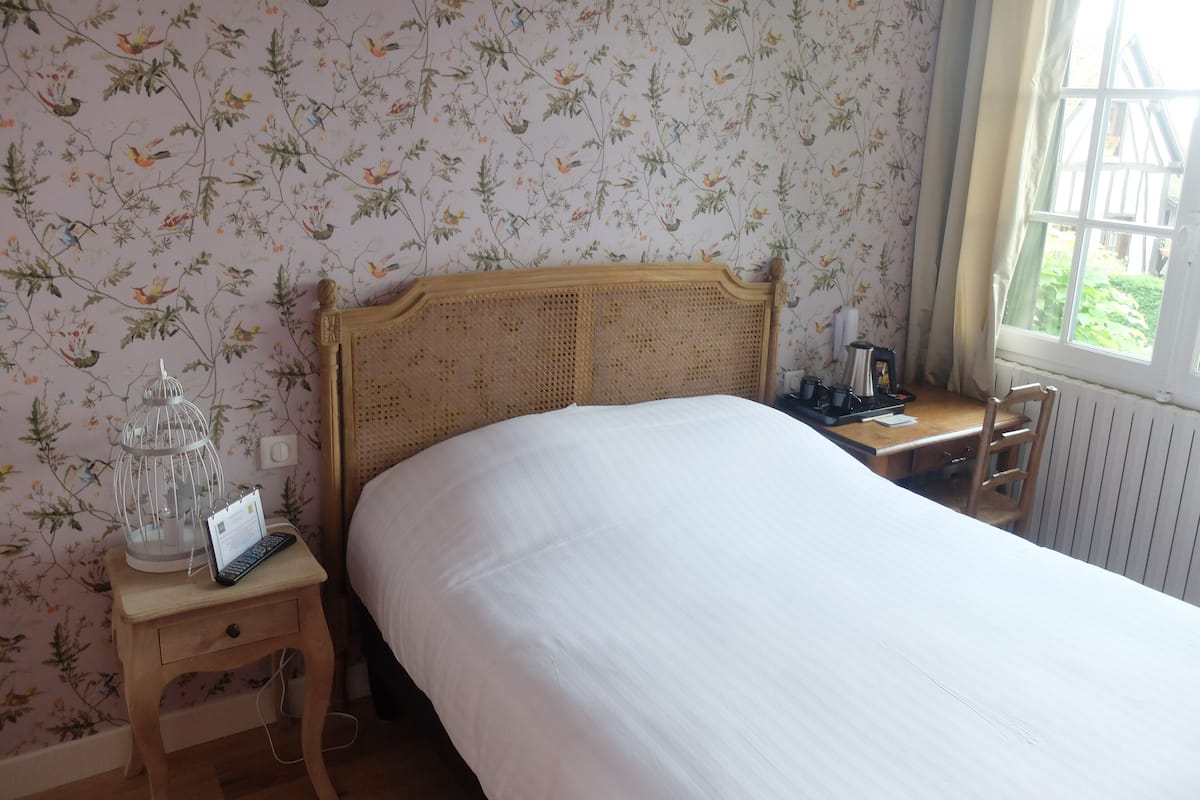 Un dormitorio con cabecera de madera, cama con ropa blanca, mesita de noche con lámpara, una silla y ventana con vista a zonas verdes.