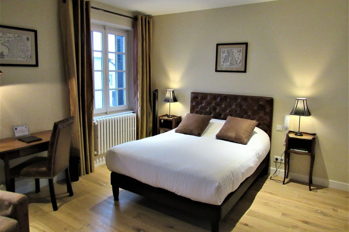 Habitación de hotel con una cama grande, dos mesitas de noche con lámparas, un escritorio con silla, un radiador y una ventana con cortinas.