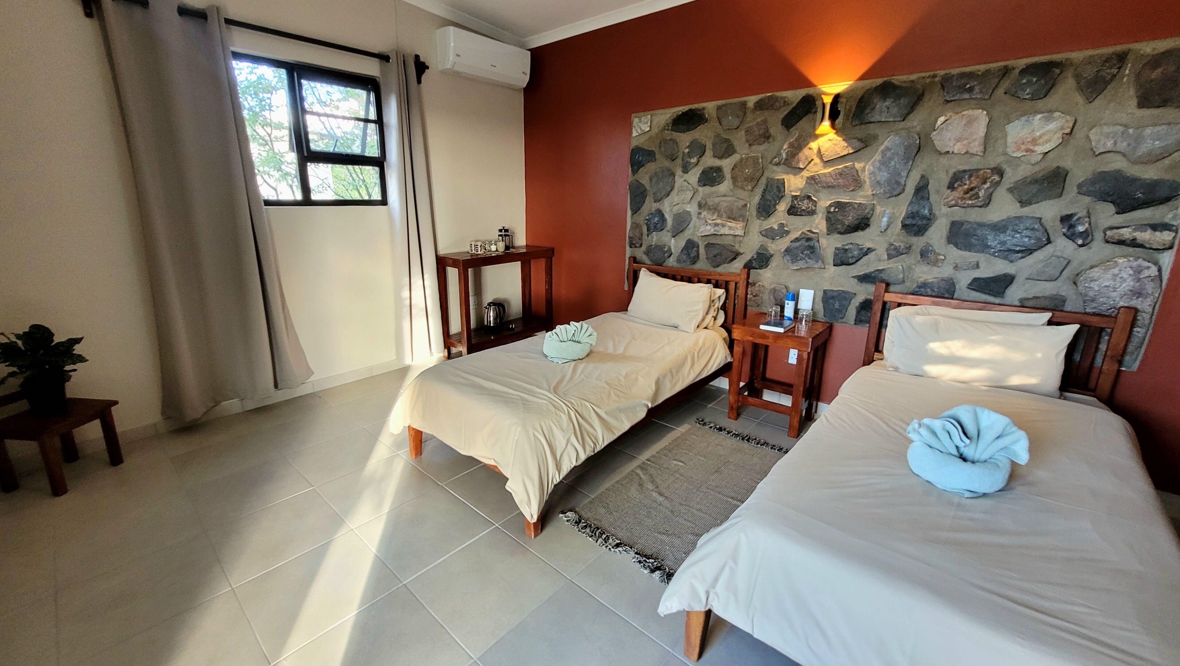 A hotel room with two beds, a stone wall, a window with curtains, and a small table with a plant.