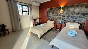 A hotel room with two beds, a stone wall, a window with curtains, and a small table with a plant. - Mondjila Safari Camp - Campsite (Outjo)