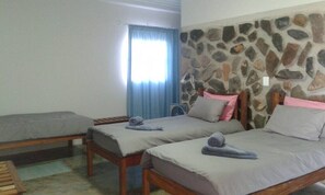 A room with two single beds, a stone wall, a window with curtains, and a wooden bench.