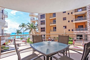 A balcony with a glass table and chairs, overlooking a beach and a building with multiple balconies.