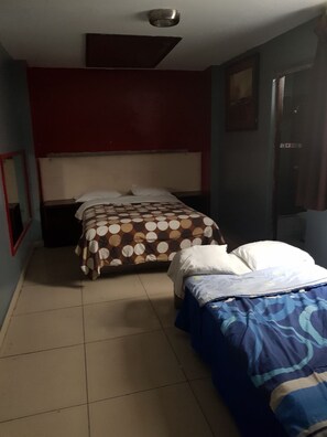Una habitación con dos camas, una pared roja, una lámpara en el techo y una puerta.
