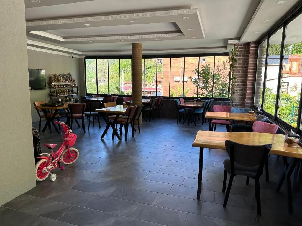 A spacious indoor dining area with multiple tables and chairs, large windows offering a view of the outdoors, and a children's bicycle in the corner.
