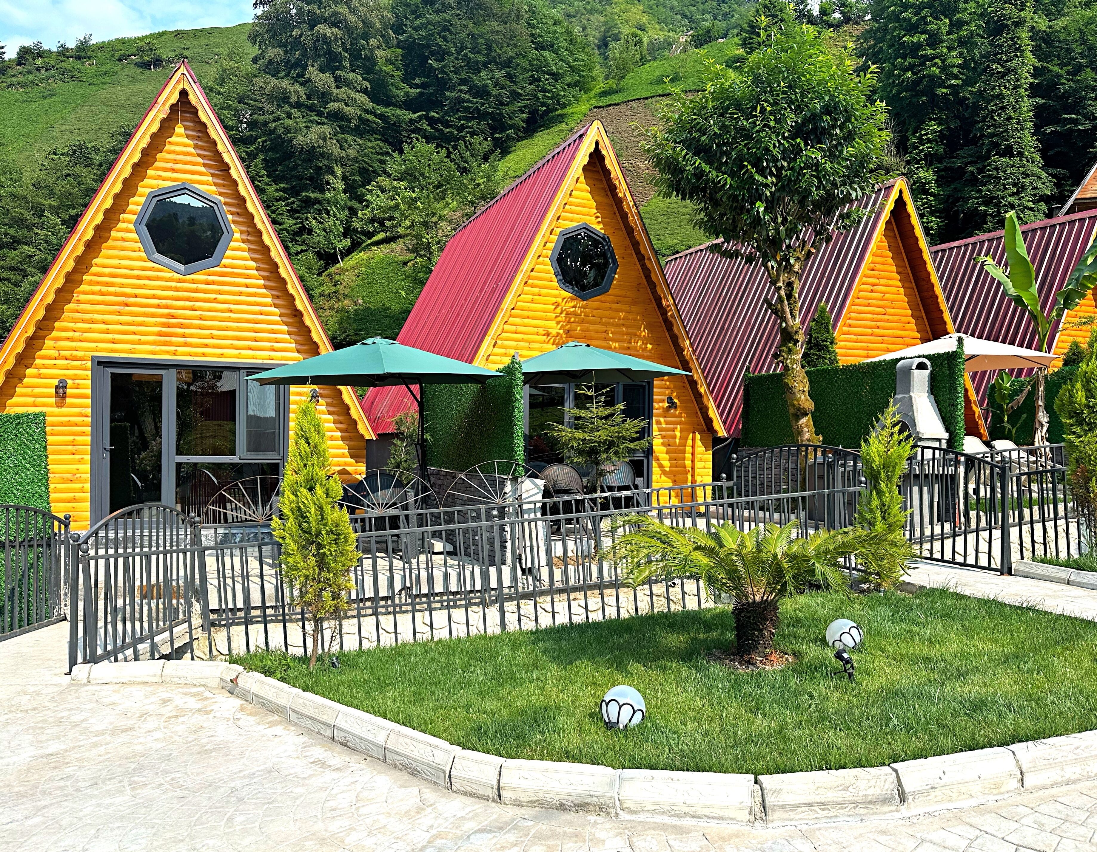 Une rangée de cabanes en rondins aux couleurs vives, dotées d’une terrasse délimitée par une clôture et d’un petit jardin.