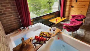 A room with a large bathtub, a wooden table with fruit and wine, and a view of a river and mountains. - Tabiat Bungalows (Camlihemsin)