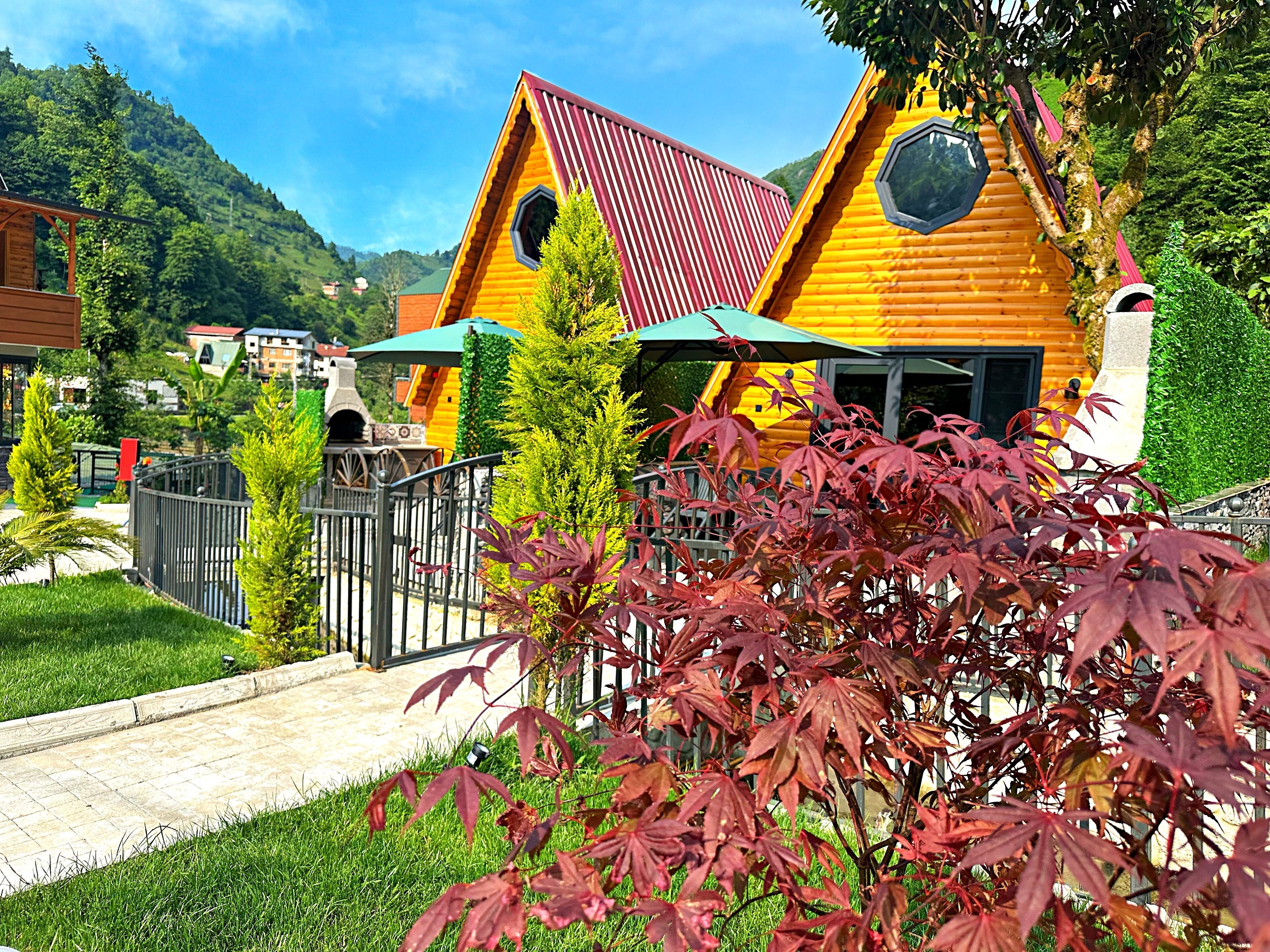 A wooden cabin with a red roof, surrounded by greenery and a paved path.