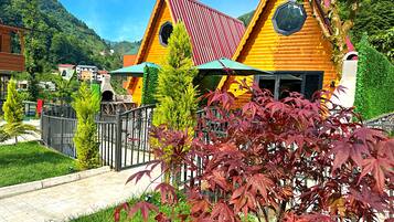 A wooden cabin with a red roof, surrounded by greenery and a paved path.
