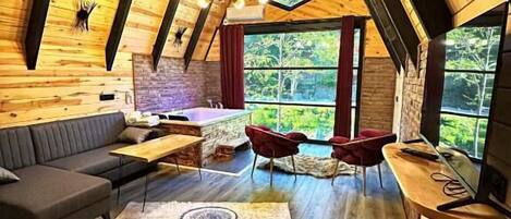 A cozy, wooden interior with a loft-style ceiling, a stone fireplace, and a large window offering a view of the outdoors.