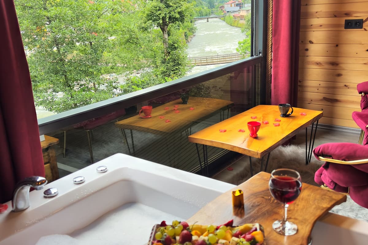 A bathroom with a bathtub, a wooden table with a fruit platter and a glass of red wine, and a view of a river and greenery outside.