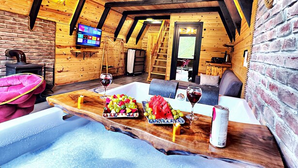 A cozy wooden cabin interior with a wooden dining table set with fruits and wine, a fireplace, and a TV on the wall. - Tabiat Bungalows (Camlihemsin)