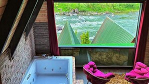 A room with a large window overlooking a river, a hot tub, and two pink chairs. - Tabiat Bungalows (Camlihemsin)