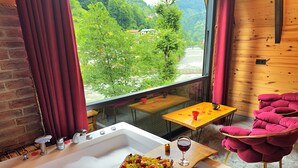 A room with a large bathtub, a wooden table with fruit and a glass of wine, a red chair, and a view of a river and greenery outside. - Tabiat Bungalows (Camlihemsin)