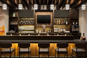 Zona de bar con una barra larga, varios estantes con botellas, vasos y un televisor de pantalla plana. Hay taburetes de bar y una máquina de café.