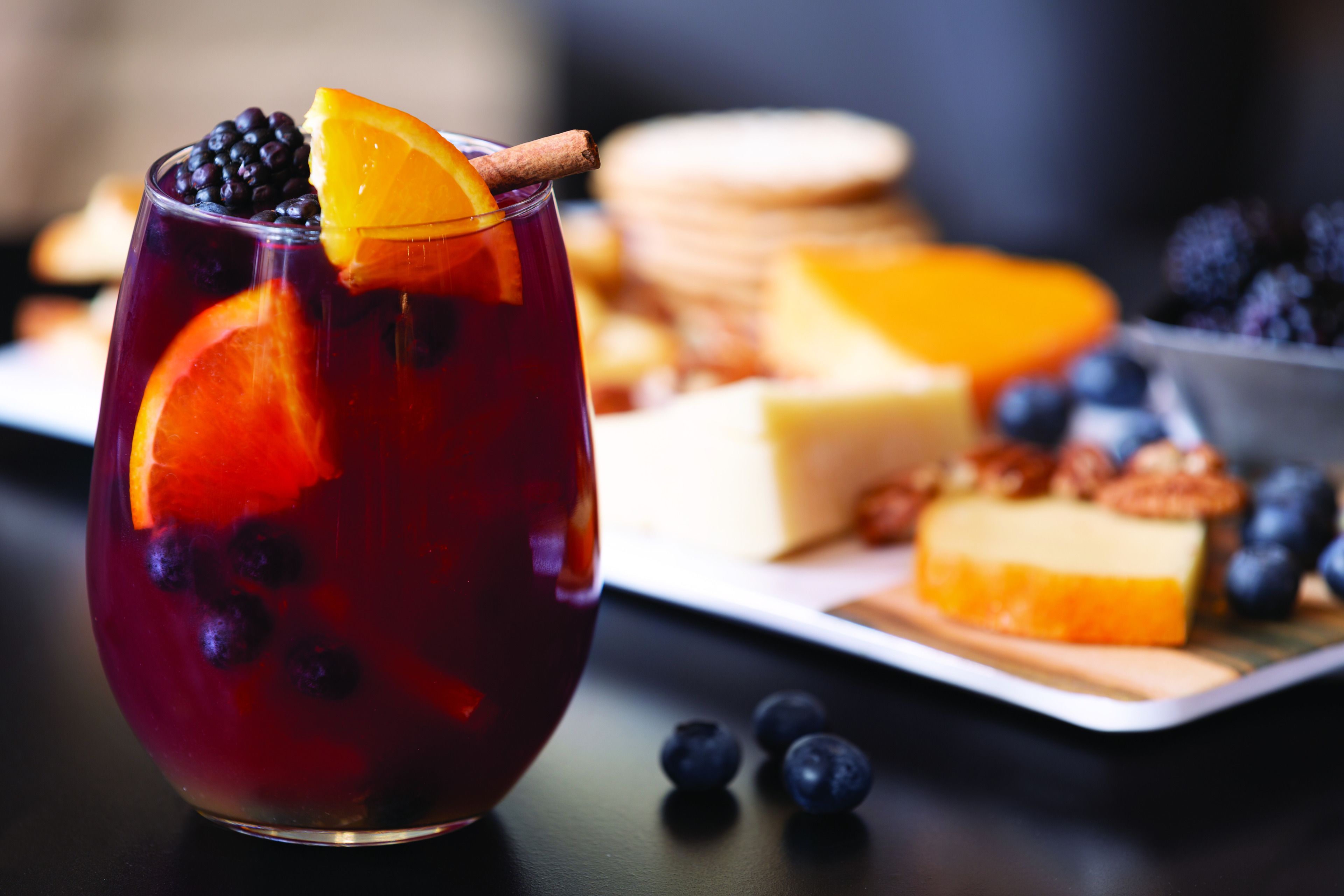 A glass of red beverage with blueberries and an orange slice, accompanied by a cheese platter with various fruits and nuts.