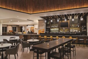 Zona de bar moderna con diversas opciones de asientos, incluyendo taburetes y mesas redondas, y un televisor en la pared.